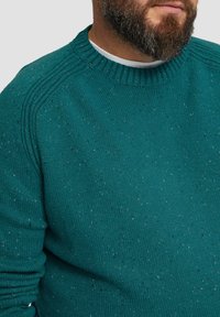 Türkis gestrickter Pullover mit rundem Ausschnitt und gerippten Details, mit gesprenkelter Textur. Die Ärmel haben eine leichte Raffung in der Nähe der Bündchen.