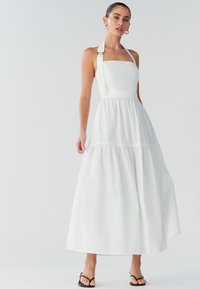 Vestido largo blanco, sin mangas, con escote cuadrado, falda fruncida y tirantes ajustables con acentos metálicos. Textura de tela suave.