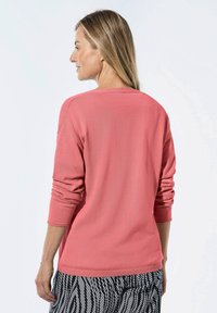 Korallenrosa Pullover mit langen Ärmeln und geripptem Saum, glatte Textur, getragen mit gemusterten schwarz-weißen Hosen. Rückansicht gezeigt.