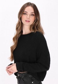Pull en maille noire à coupe décontractée, avec des manches longues et un encolure large. Porté sur un jean noir taille haute. Tissu texturé.