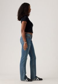 Fiatal nő félprofilban, fekete rövid ujjú felsőben, kék magas derekú farmerben, fekete sportcipőben és karkötőben, fehér háttér előtt áll.