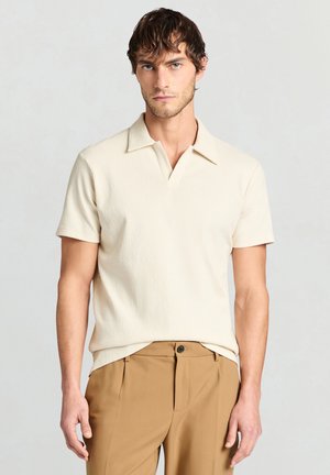 Giovane uomo con capelli castani corti, indossa una camicia crema a maniche corte con colletto e pantaloni color sabbia su uno sfondo chiaro e uniforme.