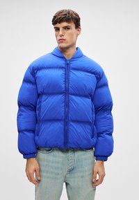 Mėlynas pūkinis striukė su užtrauktuku priekį, dėžutės formos dizainas ir elastiniai rankogaliai. Yra išsiskiriantis siuvinėjimas ir prekės ženklas ant krūtinės.