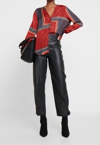 Blouse à motifs rouges et bleu marine avec manches longues et col en V, associée à un pantalon en cuir noir taille haute et des bottines noires. Sac à main noir.