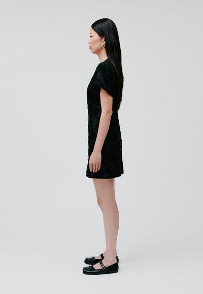 Black dress with textured floral pattern, short sleeves, fitted silhouette, above-the-knee length. Black flat shoes with buckles.