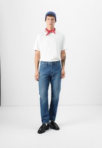Jeans de mezclilla azules, camiseta blanca, gorro azul y bandana roja. Los jeans son de corte recto; el atuendo está complementado con zapatos negros.