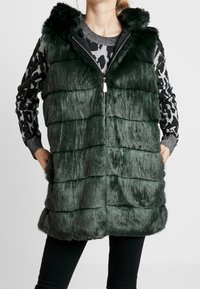 Grüne Kunstpelzweste mit Kapuze, mit horizontalen Streifen, Reißverschluss und zwei Seitentaschen. Über einem gemusterten Pullover getragen.