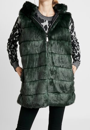 Gilet en fausse fourrure verte avec une capuche, présentant des rayures horizontales, une fermeture zippée et deux poches latérales. Porté par-dessus un pull à motifs.