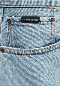 Τσέπη από ανοιχτό μπλε τζιν με πορτοκαλί ραφές, δύο μεταλλικά καρφιά και μια μαύρη ετικέτα Quiksilver ραμμένη πάνω από την μικρότερη τσέπη για κέρματα.