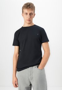 Ung mann med kort hår iført en ensfarget svart kortermet t-skjorte og lysegrå jeans, med den ene hånden i lomma, mot en hvit bakgrunn.