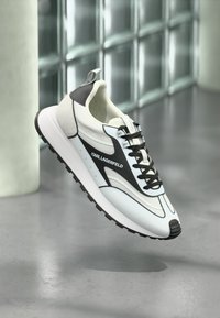 White and black athletic sneaker with a textured surface, featuring a round toe, laces, and a cushioned sole. Karl Lagerfeld logo displayed prominently.