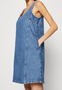 Denimmekko, jossa on olkaimeton muotoilu, V-pääntie, sivutaskut ja väljä muoto. Kankaan väri on keskisininen ja siinä on näkyvät saumur stitching.