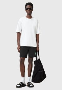 Camiseta blanca de gran tamaño, pantalones cortos negros con un logo verde, sandalias negras, calcetines blancos y una bolsa de mano negra con texto en negrita.