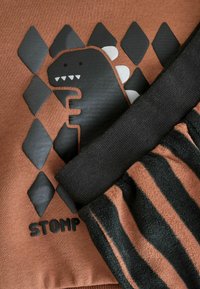 Hnedá mikina so čiernou grafikou dinosaura a slovom "STOMP", spárovaná s čiernymi a hnedými pruhovanými flísovými nohavicami.