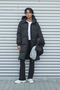 Melns pūkains mētelis ar kapuci un sānu kabatām, kopā ar baltu augšu, melnām biksēm un baltiem sporta apaviem; rokās pelēka aksesuāru soma.