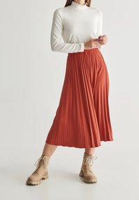 Langer, plissierter Rock in rostfarben, kombiniert mit einem weißen Rollkragenpullover und hellbraunen Schnürstiefeletten. Weicher Stoff mit fließendem Design.