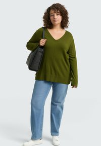 Olivgrüner V-Ausschnitt-Pullover mit langen Ärmeln, lockere Passform; kombiniert mit hellblauen Straight-Leg-Jeans und weißen Sneakern, während sie eine schwarze Tasche trägt.