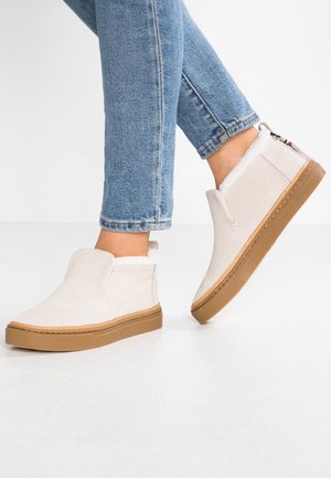 Stopy w beżowych zamszowych botkach typu slip-on z brązowymi gumowymi podeszwami i niebieskich jeansach na tle jednolitej białej ściany.