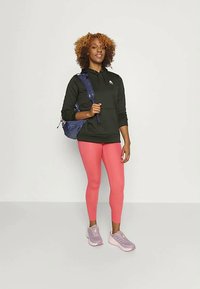 Γυναίκα φορώντας μαύρη κουκούλα, κοραλί κολάν, ροζ αθλητικά παπούτσια και μπλε σακίδιο, στέκεται μπροστά σε απλό ανοιχτό γκρι φόντο.