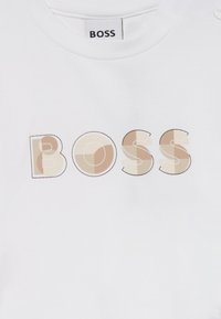 Camiseta de algodón blanca con un logo "BOSS" en patrones geométricos beige y marrón. Diseño clásico de cuello redondo con una textura suave.