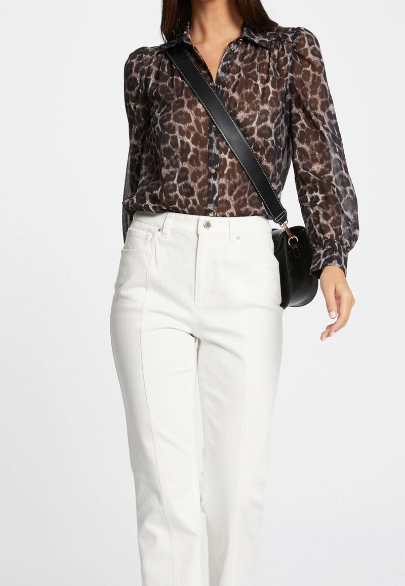 Femme portant un chemisier transparent à manches longues imprimé léopard, un pantalon blanc taille haute et un sac bandoulière noir avec une sangle large.