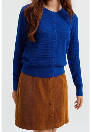 Blauwe gestructureerde gebreide cardigan met ronde hals en knoopsluiting, gecombineerd met een bruine korduroy rok met een knoopsluiting aan de voorkant.