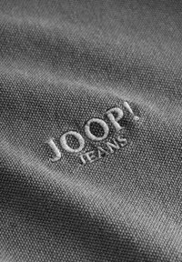 Tissu gris affichant le logo brodé "JOOP! JEANS" en blanc, avec un motif de tissage texturé et un léger éclat.