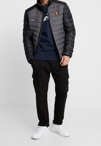 Chaqueta acolchada negra y gris con cuello alto, que presenta un logo en el pecho, superpuesta sobre un suéter azul marino y combinada con pantalones de carga negros.