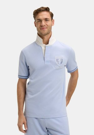 Homme portant un polo bleu clair à manches courtes avec col blanc et logo, debout devant un fond clair uni, souriant légèrement.
