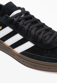 Zapatillas de ante negro con franjas de cuero blanco, cordones negros y suela de goma texturizada marrón. Presenta un logo dorado y costuras en tono similar.