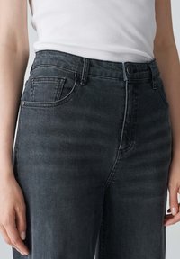 Jeans in denim grigio scuro con una texture liscia, dotati di chiusura a bottone, passanti per la cintura e tasche frontali. Design a gamba dritta, con dettagli minimi.