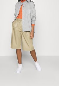 Ljusgrå skjortjacka över en orange ribbad polotröja, kombinerad med beige veckade shorts och vita höga sneakers.