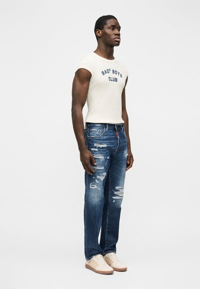 Homme debout portant un débardeur blanc avec le texte « BAD BOYS CLUB », un jean bleu déchiré et des baskets beiges sur un fond blanc.