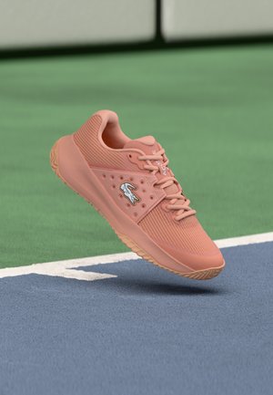 Rosa Lacoste tennissko med lisser og logo svever over en blå og grønn tennisbane nær den hvite grensen.