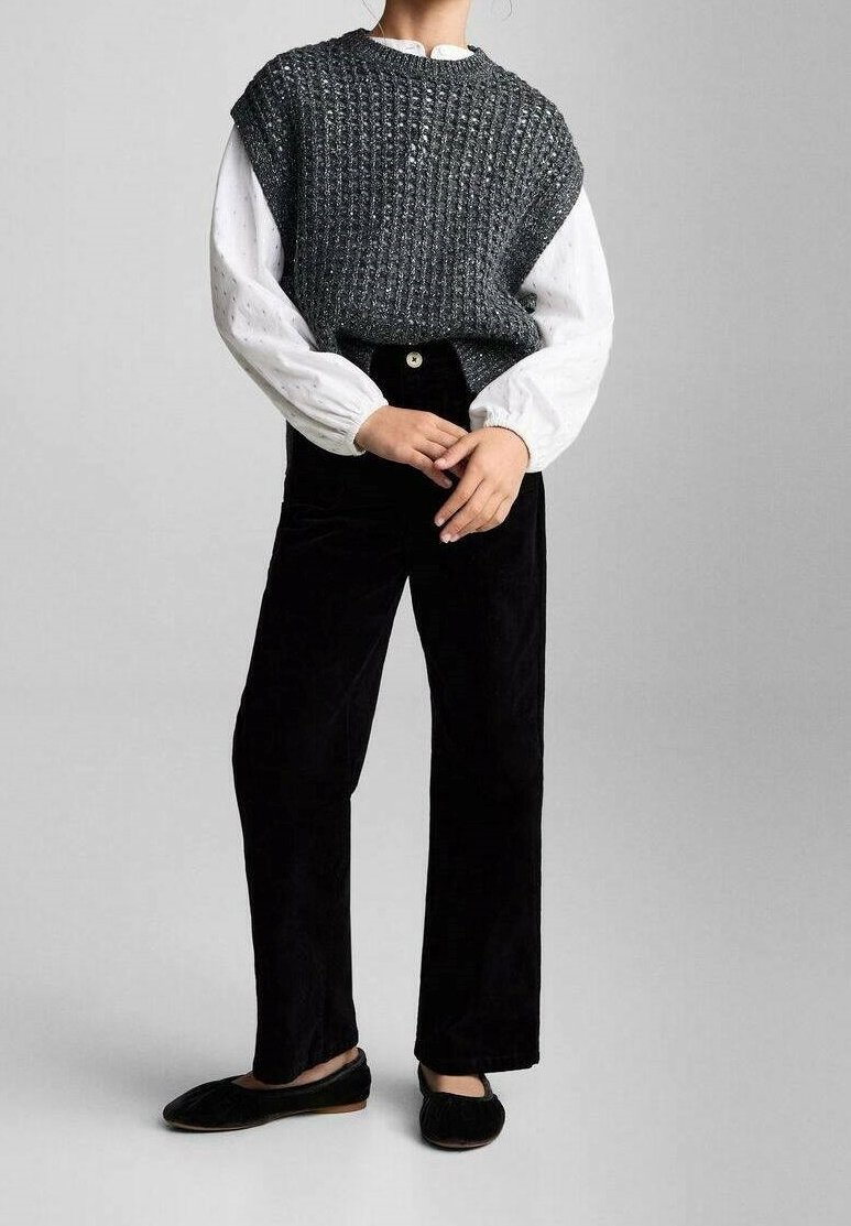 Personne portant un pull sans manches gris foncé texturé sur une blouse blanche à manches longues, un pantalon noir et des chaussures plates noires, debout, les mains jointes.