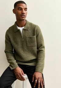 Suéter de color verde oliva con cuello con cremallera, tejido texturizado y un bolsillo en el pecho, combinado con pantalones negros. Apto para uso casual.