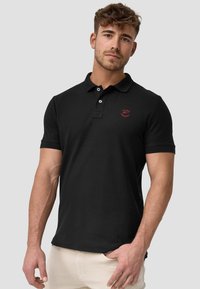 Sort polo-shirt i bomuld med korte ærmer, der har en lukning med to knapper og et lille rødt logo på venstre bryst. Glat tekstur.