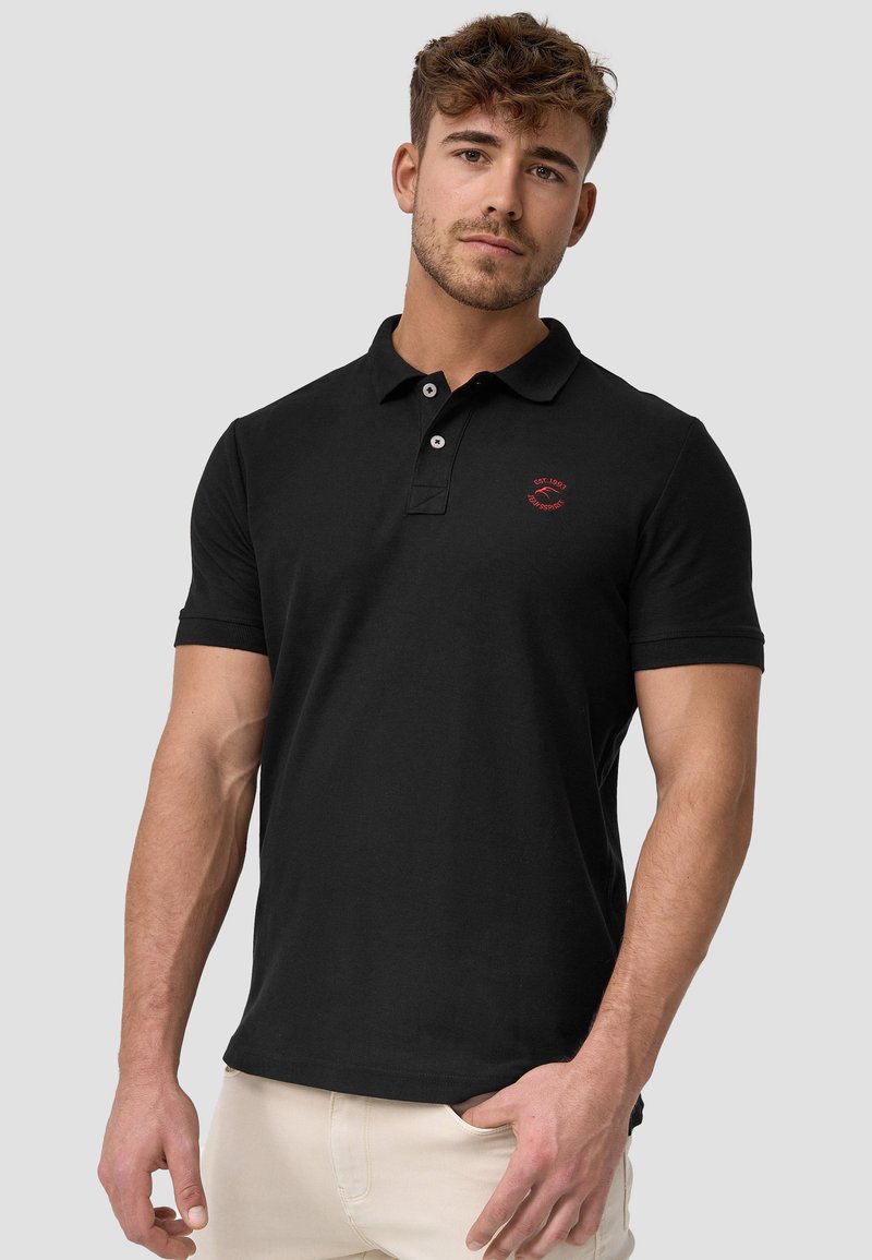 Sort polo-shirt i bomuld med korte ærmer, der har en lukning med to knapper og et lille rødt logo på venstre bryst. Glat tekstur.
