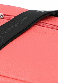 Sintetična torba koralne roza barve z teksturirano površino, opremljena z črnim trakom z blagovno znamko "TOM TAILOR" in vidnim kovinskim zadrgo.