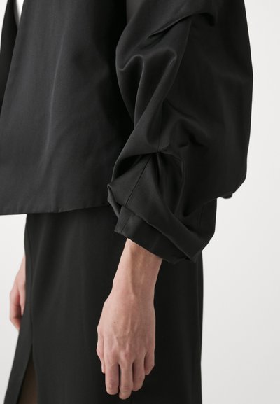 Black fabric top with oversized, draped sleeves and cuff detailing. The design features a cropped silhouette, paired with sleek black skirt.