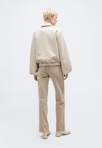 Persoană cu păr blond scurt, văzută din spate, purtând o jachetă bej, pantaloni drepti asortați și pantofi bej cu toc bloc, pe fundal alb.