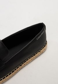 Juoda espadrilė su lygiu odiniu viršumi ir šiaudiniu padu. Yra subtilus prekės ženklas "Calvin Klein" šone.