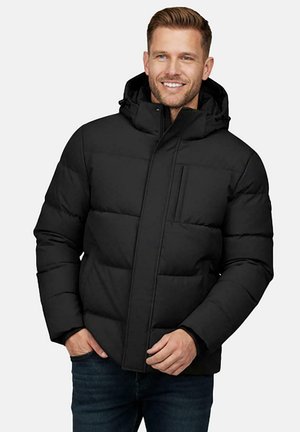 Man wearing a black padded winter jacket with a hood, smiling, hands partially in pockets, standing against a plain background.