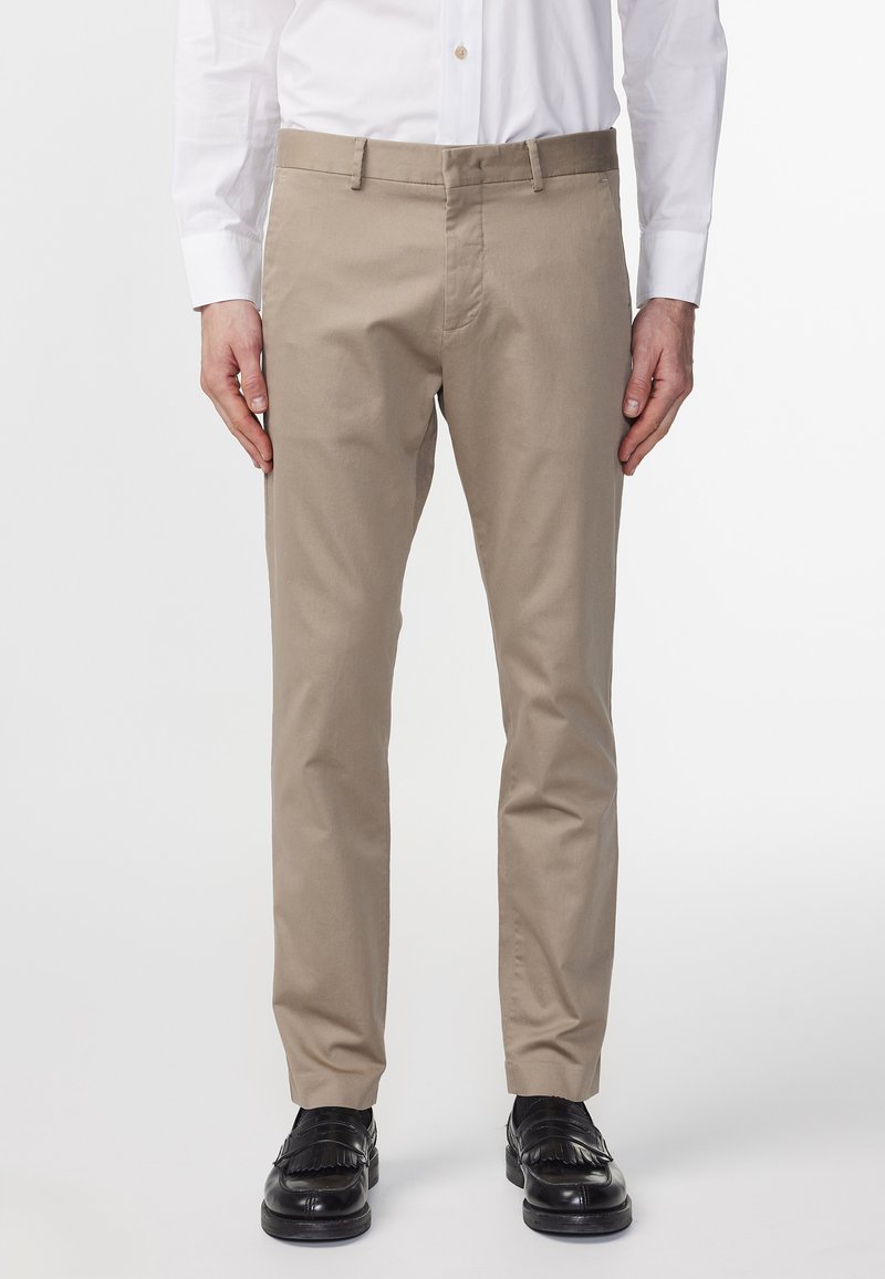 Beige chinos gjorda av bomull med rak bendesign. Har en midjeband med bältesömmar och sidofickor, tillsammans med svarta skor.