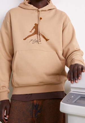 Person trägt beigen Hoodie mit braunem Jumpman-Logo und dem Schriftzug "Flight" und berührt eine weiße Bürogerät, Ansicht von Torso und Händen.
