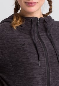 Mørkegrå zip-up hoodie lavet af tekstureret stof; har en hætte med snor og broderet logo. Lynlåsen og lommerne er ikke synlige.