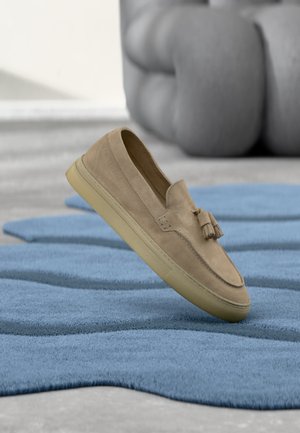 Beige suède loafer met tassels zwevend boven een blauw gestructureerd vloerkleed, zachte grijze zitplaats op de wazige achtergrond.