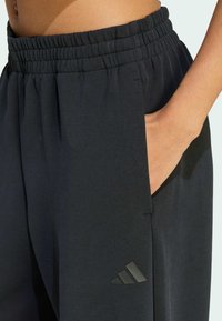 Ruka odmara u džepu crnih Adidas trenirki s elastičnim pojasom i logotipom blizu bokova.