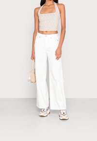 Kvinna i beige ärmlös crop top, vida vita byxor, vita och beige sneakers, håller en beige handväska vid sidan.