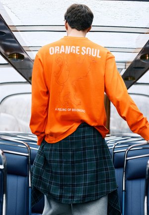 Persoon in een feloranje trui met de tekst "ORANGE SOUL" en een leeuwengrafiek, staand in een bus met blauwe stoelen en glazen dak.